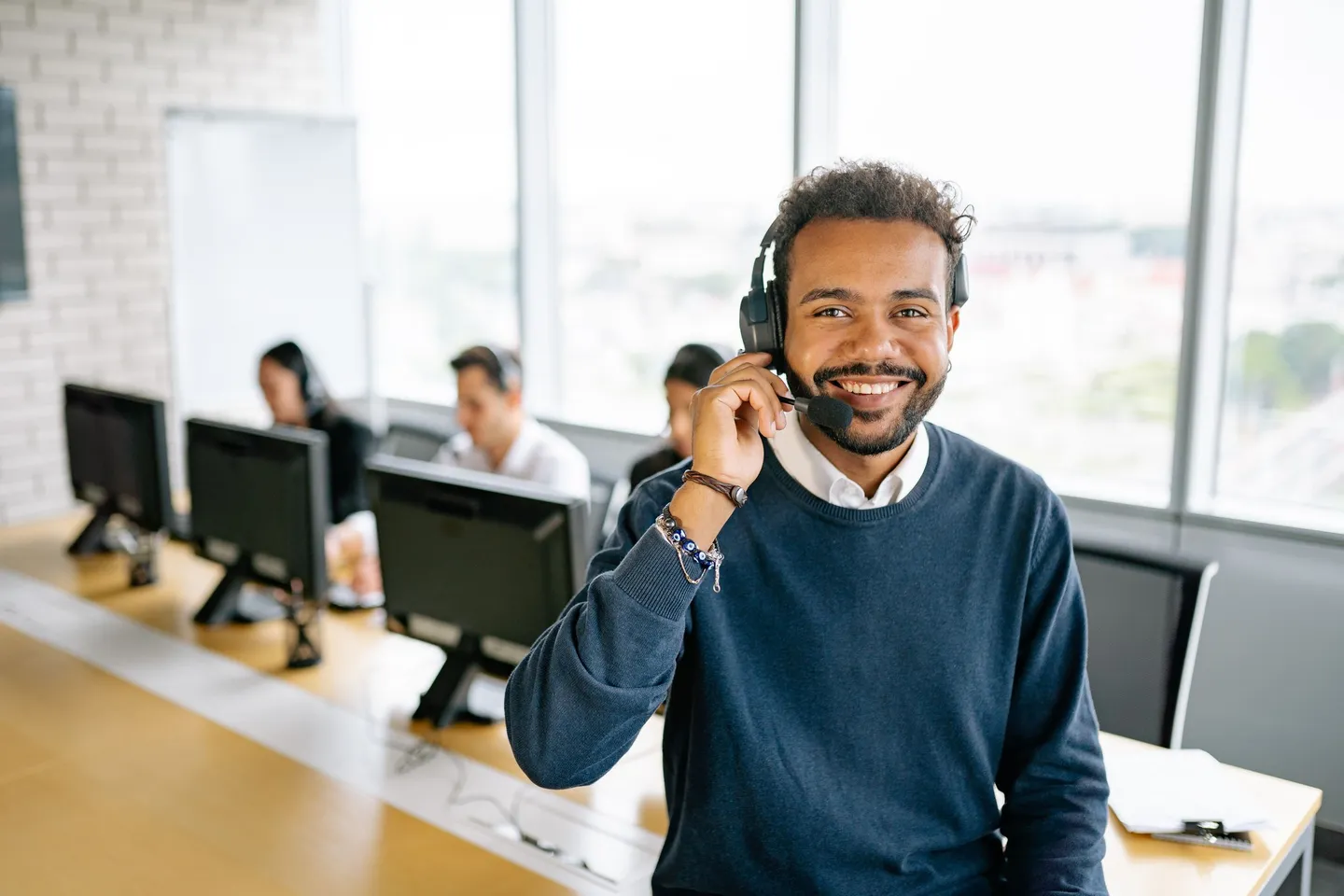 Smiling sales rep wearing a headset in an open-plan office, with colleagues working at monitors behind him