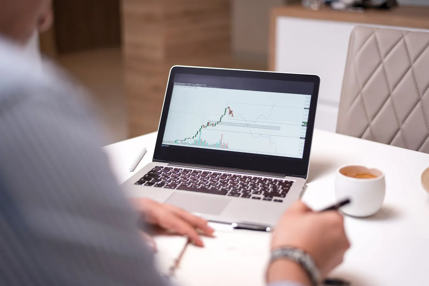 Person with a pen reviewing a chart on a laptop at a white desk, next to a cup of coffee