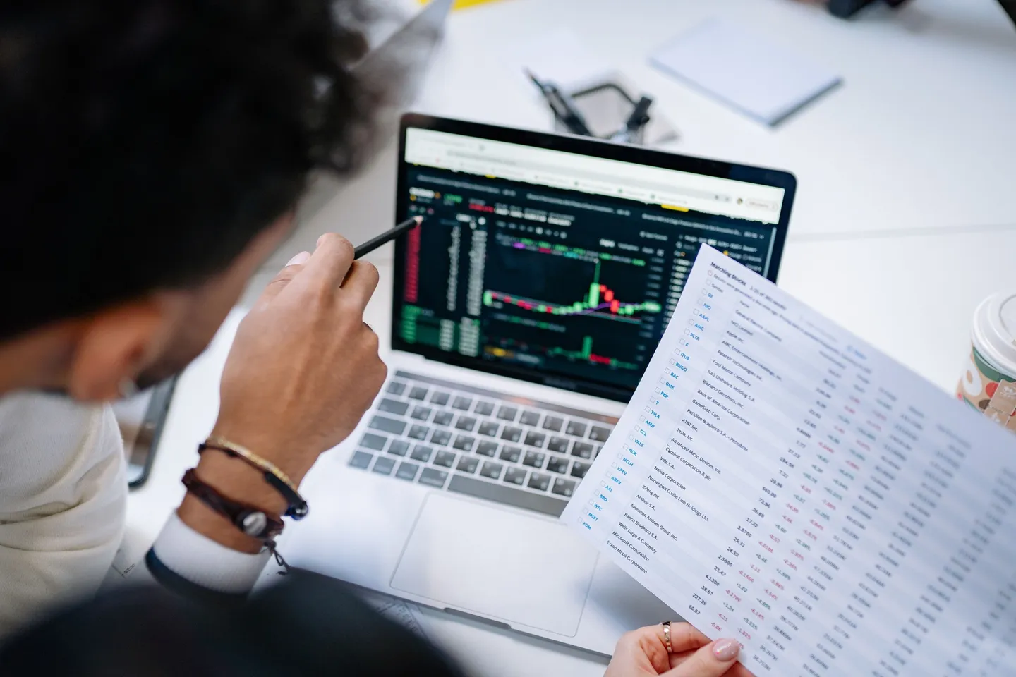 Person pointing a pen at a trading chart on a laptop while another holds a printed table of figures