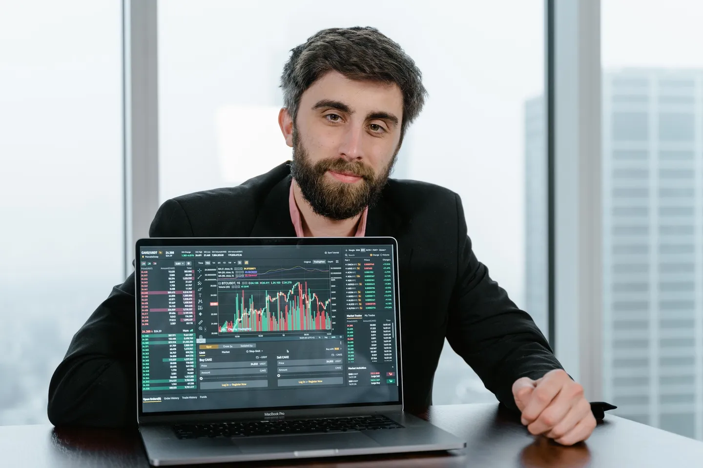 Bearded man in a dark blazer seated behind a laptop that shows a trading dashboard with price charts